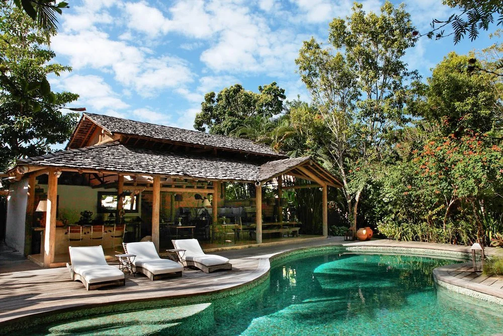 A backyard with a pool, outdoor lounge chairs, a shaded wooden structure, surrounded by lush trees and blue sky.