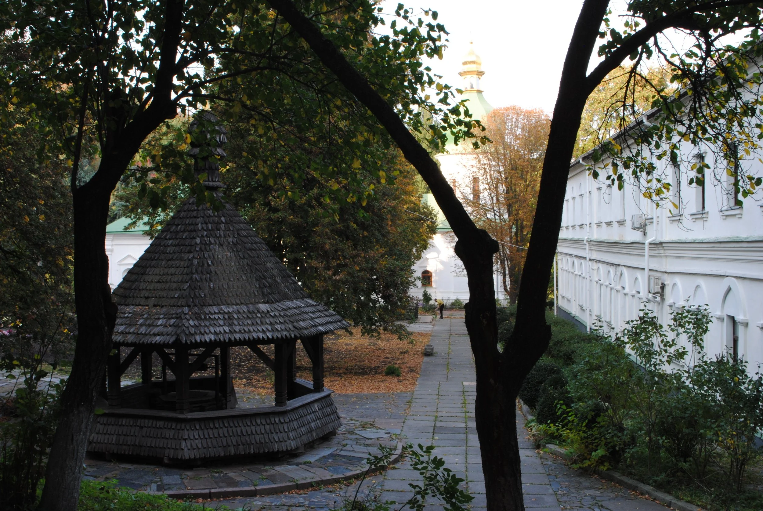 A peaceful courtyard surrounded by white buildings, with a small wooden gazebo and tall trees with autumn leaves. A person walks along a paved pathway in the background.