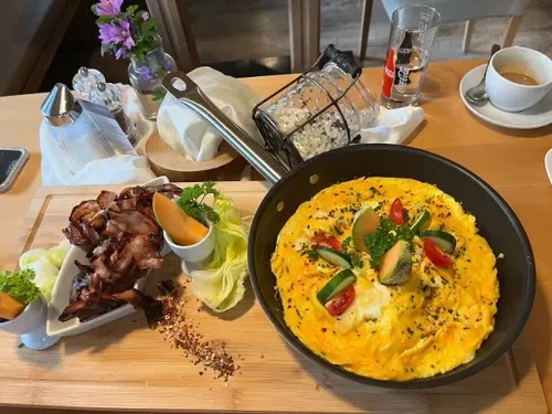 Fried eggs with vegetables in a black skillet, crispy bacon, and garnishes on a wooden tray with a side of lettuce, orange slices, condiments, a glass of water, a cup of coffee, and a small vase with purple flowers on a restaurant table.