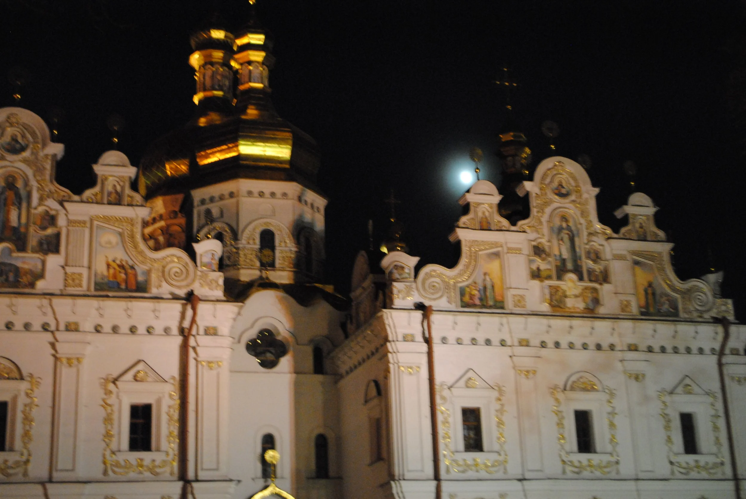 Night view of an ornate church with onion domes, illuminated against the dark sky. The moon is visible behind the church.