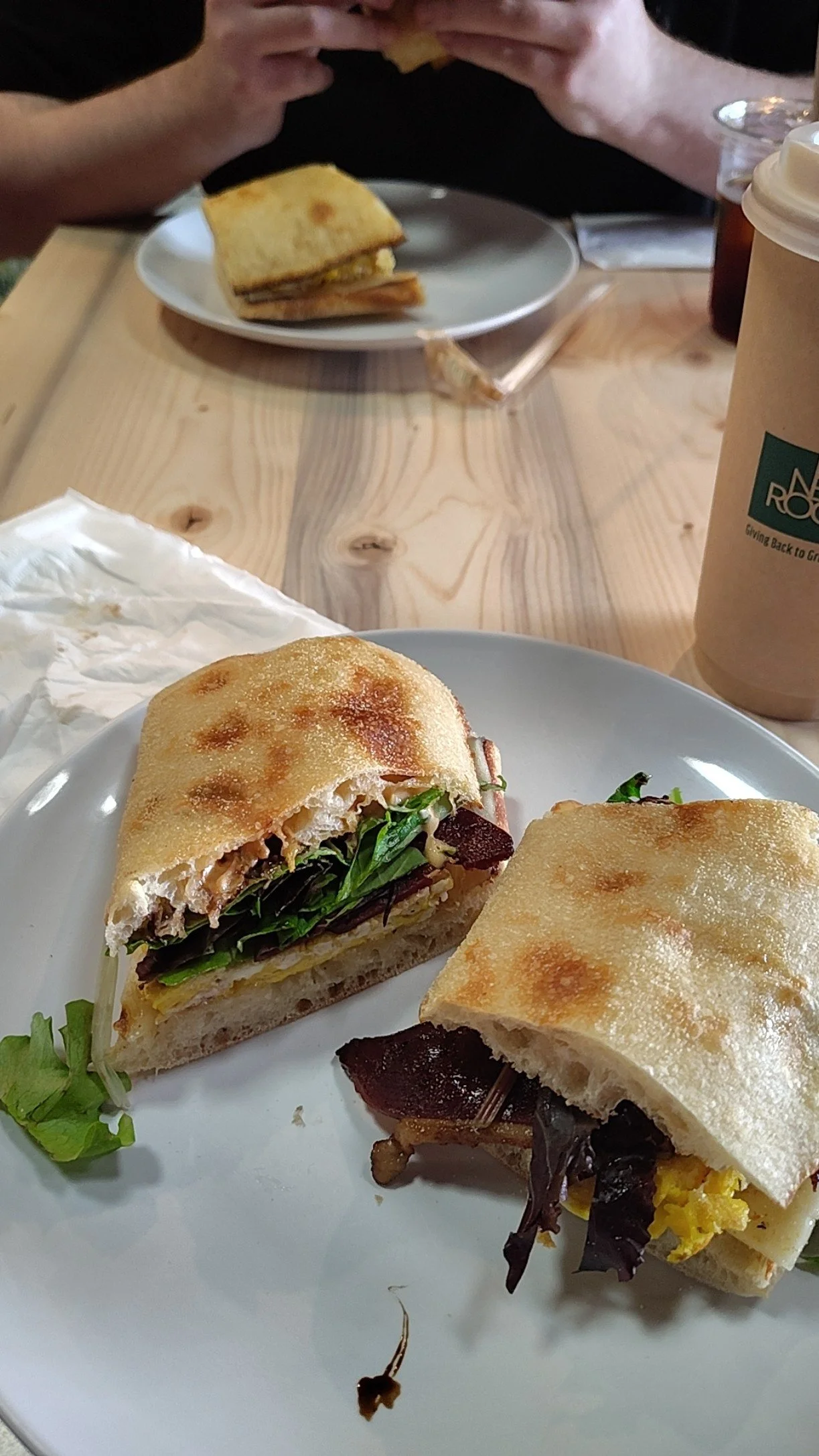 Close-up of a white plate with two sandwiches filled with greens, cheese, and meats on a wooden table, with a text on a cup in the background.