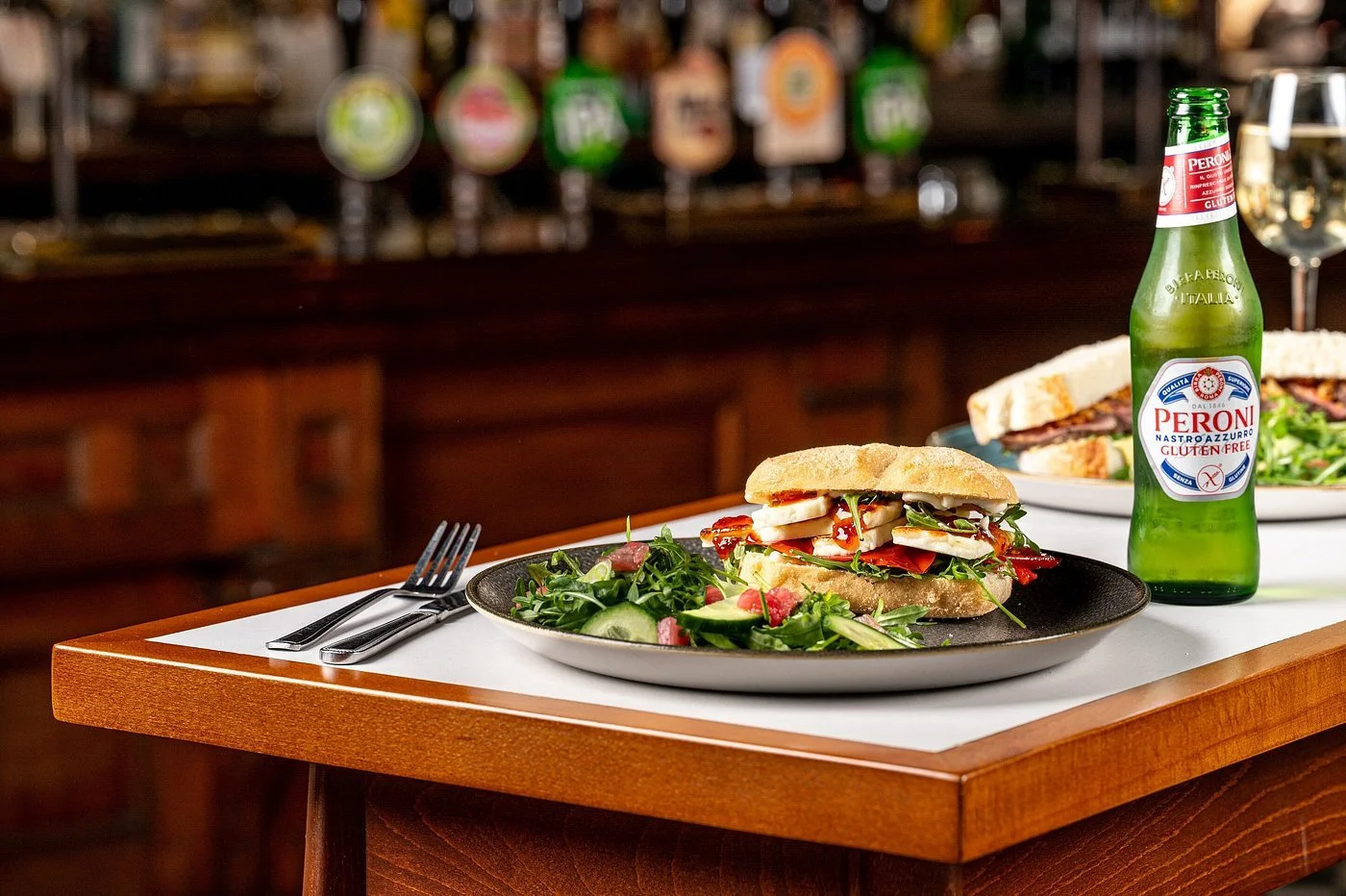 A sandwich on a black plate with a side salad, a fork and knife, a partially visible sandwich in the background, and a bottle of Peroni gluten-free beer on a wooden table in a restaurant setting.