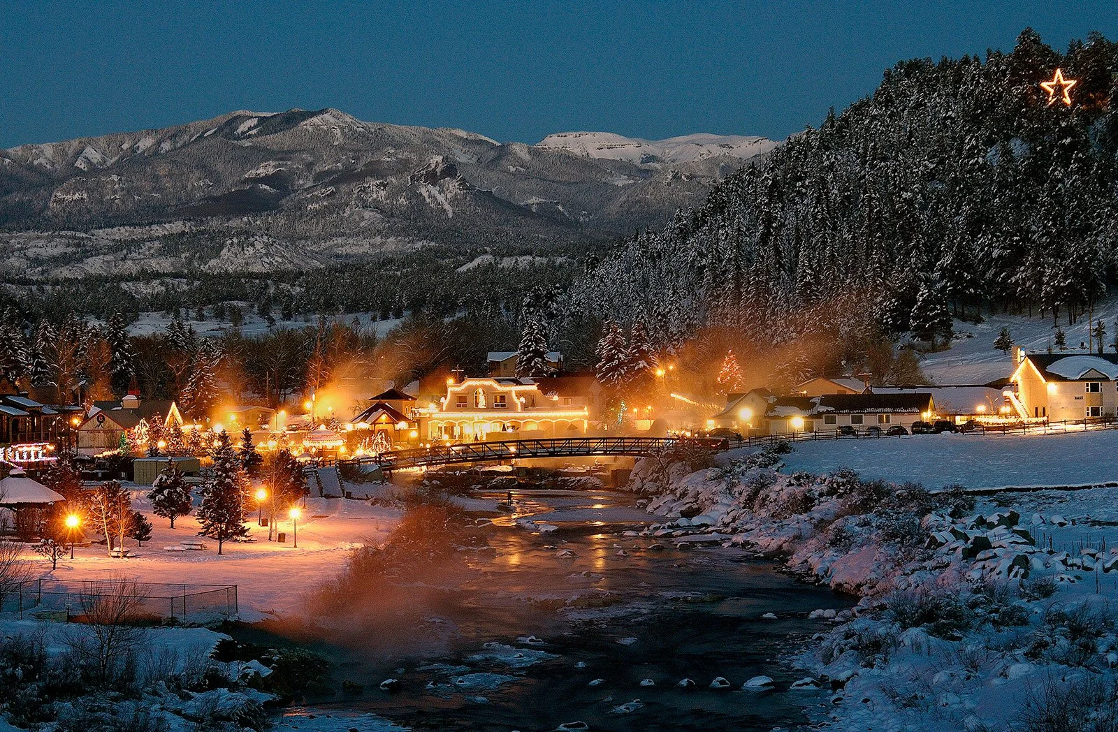 A snowy mountain town at dusk with houses decorated with Christmas lights, a river flowing through the town, and snow-covered trees and mountains in the background, with a glowing star in the sky.