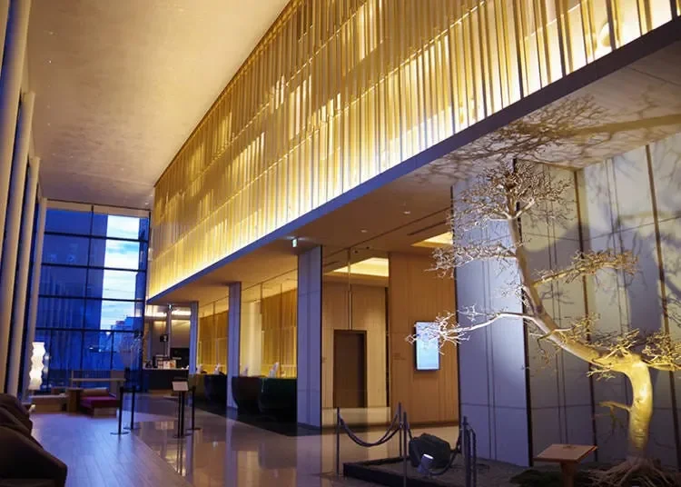 Interior of a modern hotel lobby with a large illuminated golden railing, a tall window revealing a cityscape, and a decorative tree in the corner.