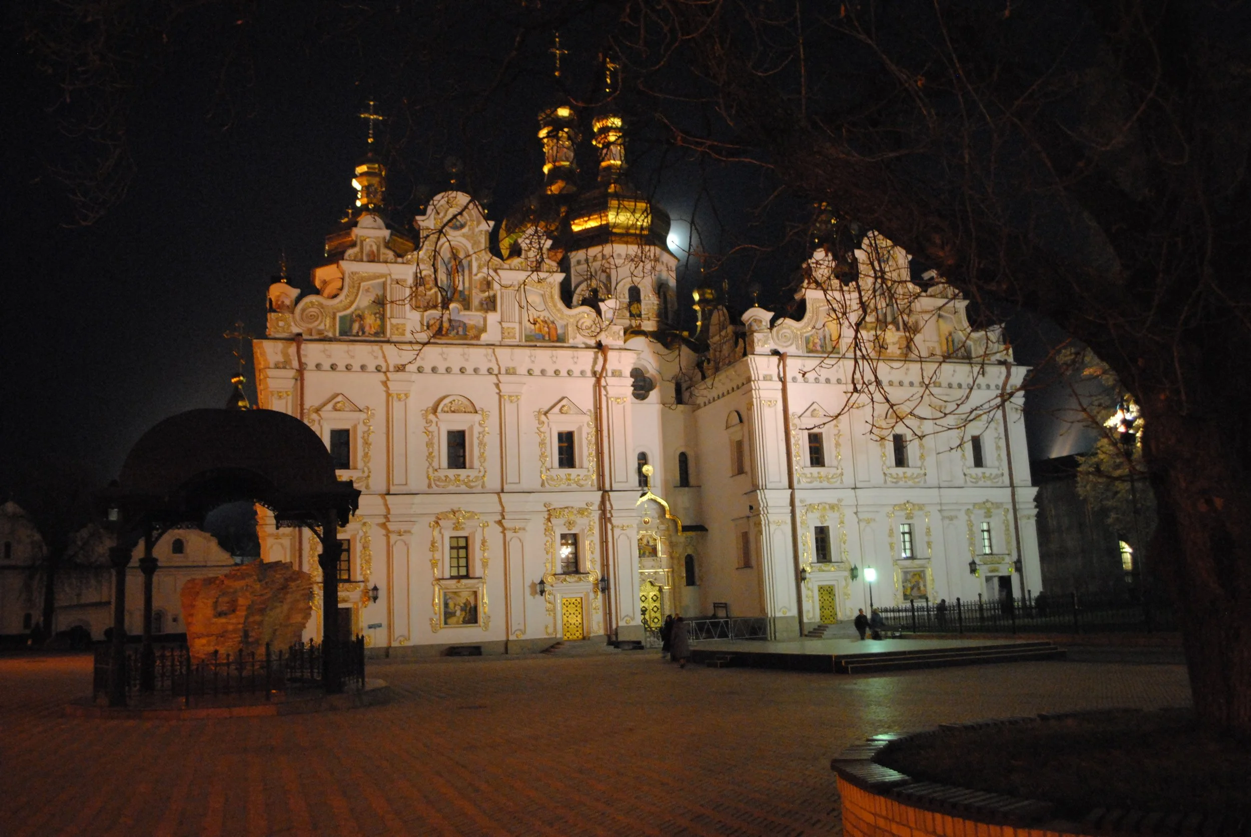 Night scene of a decorated church with ornate white and gold architecture, illuminated against the dark sky, with leafless trees in the foreground.