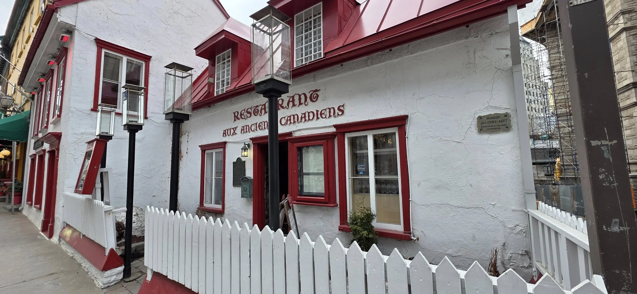 Exterior of a restaurant with white walls and red window frames, red trim, and a sign that reads "Restaurant aux Ancients Canadiens." There are black lampposts and a white picket fence along the sidewalk, with construction scaffolding visible on the 