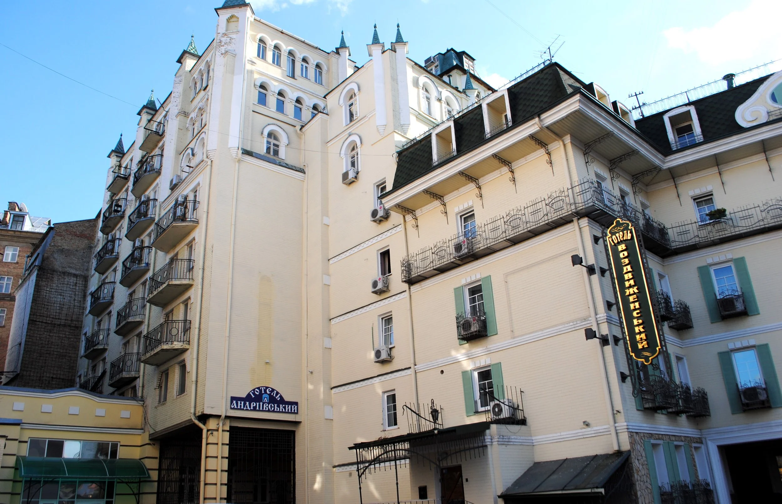A multi-story building with white walls, various windows, and balconies with black railings. The building features pointed towers and a sign in a foreign language, likely Cyrillic, indicating it may be a hotel or commercial property. The sky is clear