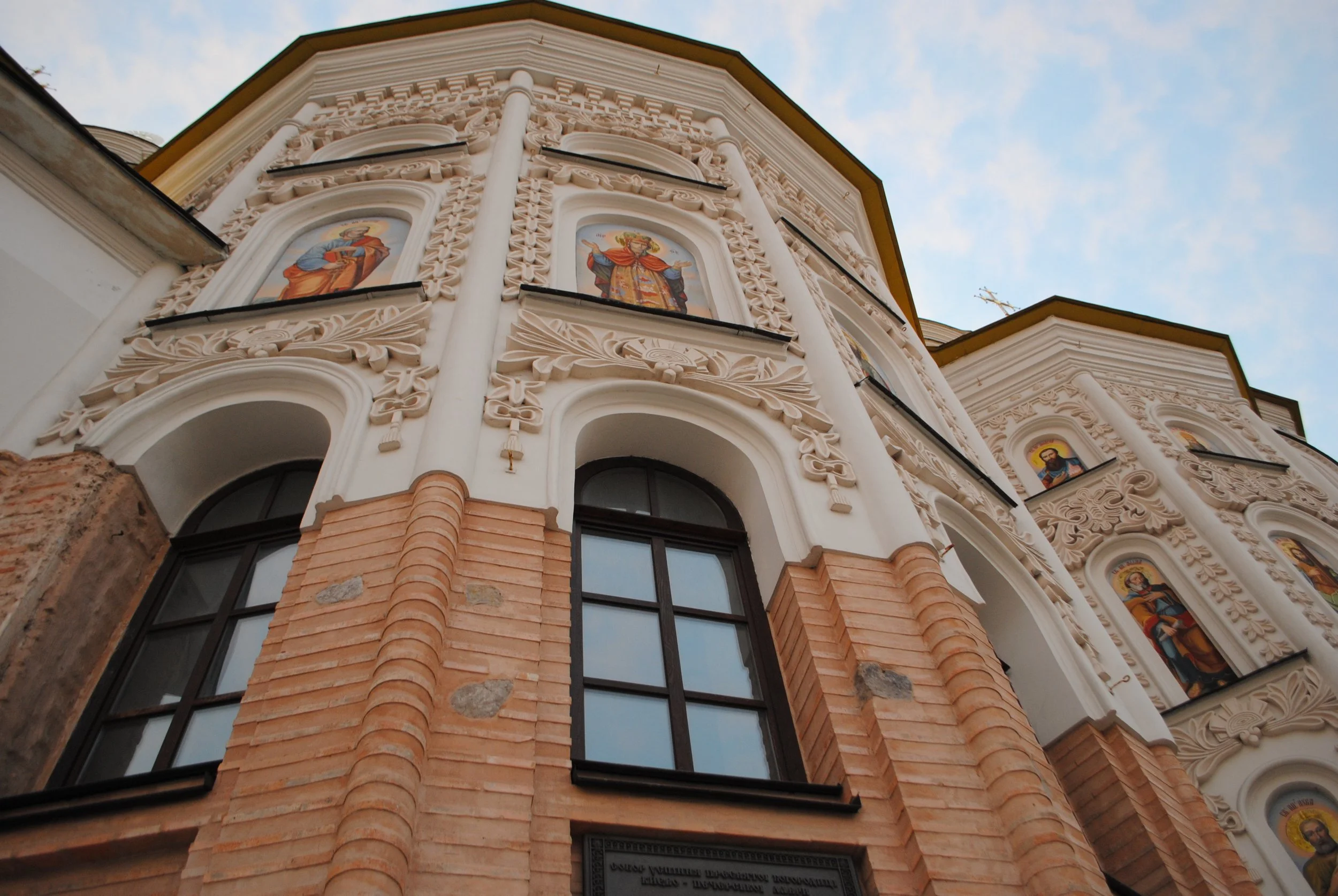 View of a church facade with intricate white stucco decorations, colorful religious icons in arched niches, large arched windows, and brick accents at the base, taken from a low angle against a blue sky
