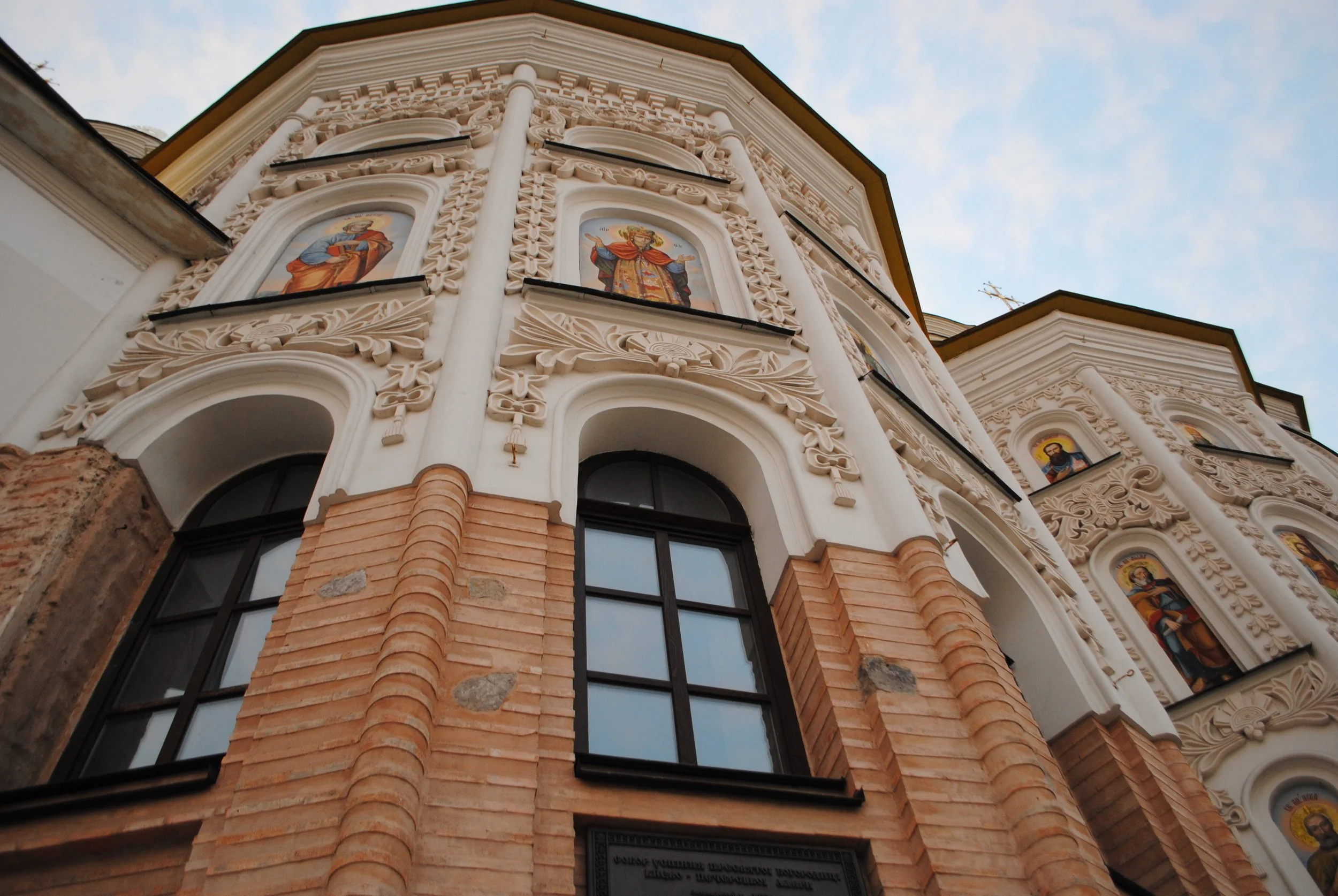 Low-angle view of a church facade with ornate white stucco and brickwork, featuring religious icons in arched niches against a partly cloudy sky.