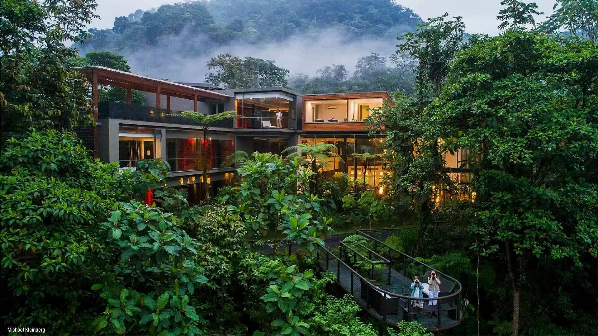 Luxury house with large glass windows surrounded by lush green trees and mountains in the background, with two women standing on a small bridge in the foreground.