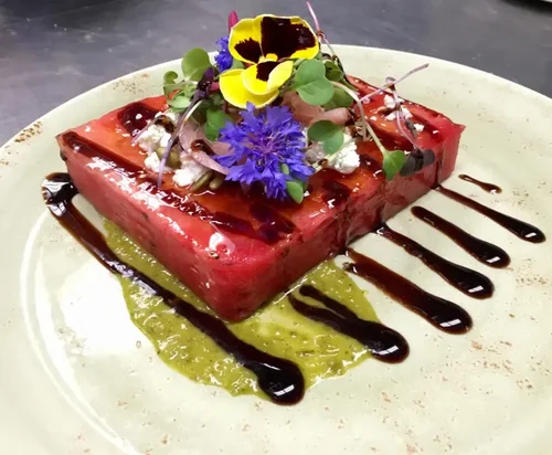 A plated dessert featuring a red, square-shaped mousse garnished with edible flowers, microgreens, and a drizzle of chocolate sauce on top and around, served on a light-colored plate.