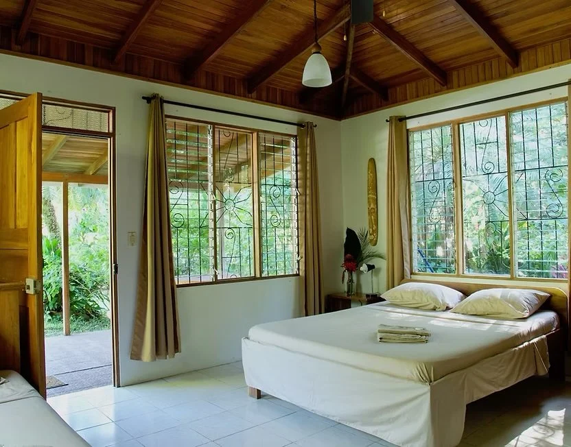 A cozy bedroom with a wooden ceiling, large windows with iron grills, a bed with white linens and pillows, a bedside table with flowers, and an open door leading outside to lush greenery.