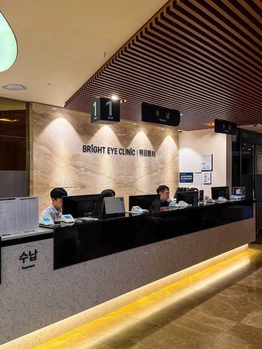 Reception area of Bright Eye Clinic with four receptionists seated behind a counter, several computer monitors, and payment signs in Korean and English.
