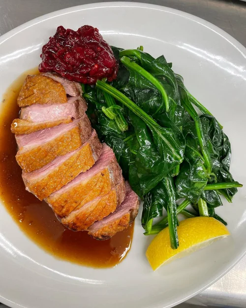 Sliced duck breast with crispy skin, topped with berry compote, served with cooked spinach, a lemon wedge, and a side of dark sauce on a white plate.