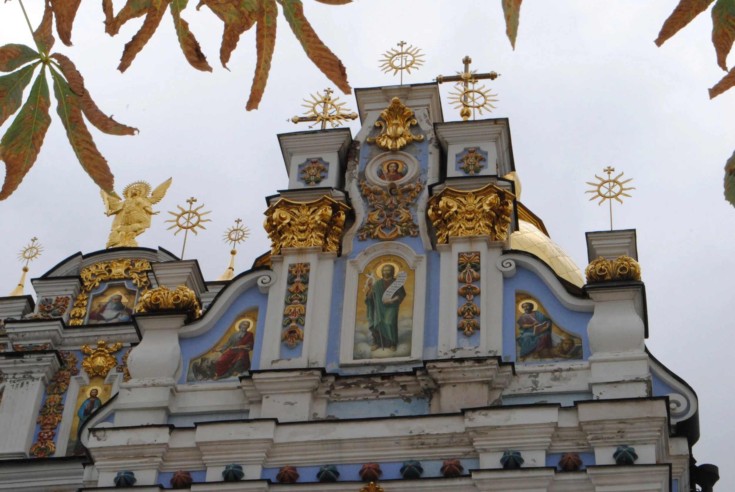 The top of a church or cathedral with ornate architectural details, gold accents, religious icons, and decorative crosses on top, viewed from below against a cloudy sky.