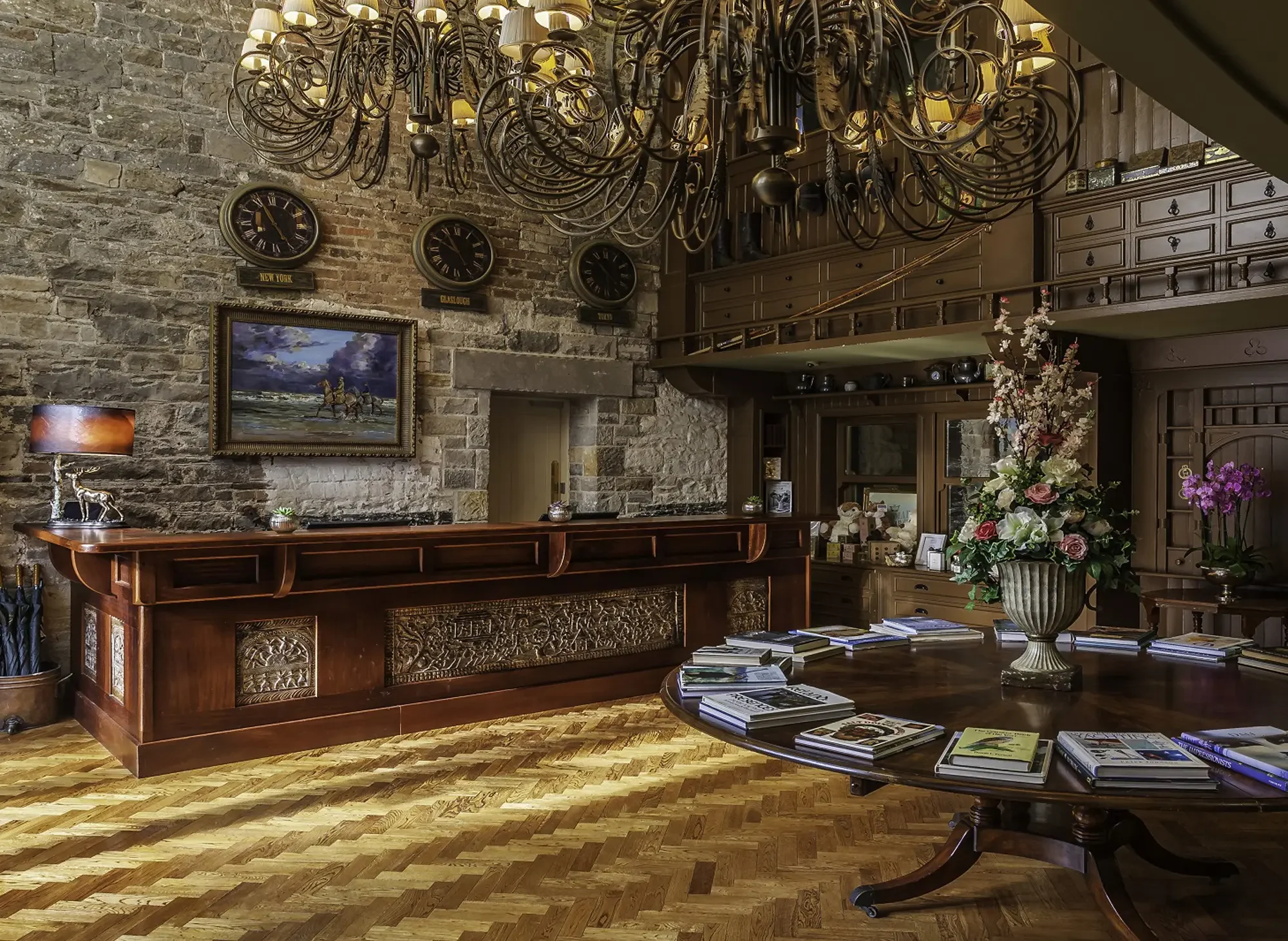 A cozy hotel lobby with a stone brick wall, a large ornate wooden reception desk, a chandelier, various clocks showing different time zones, framed artwork, a seating area with a round table with books, a flower arrangement, and decorative items.