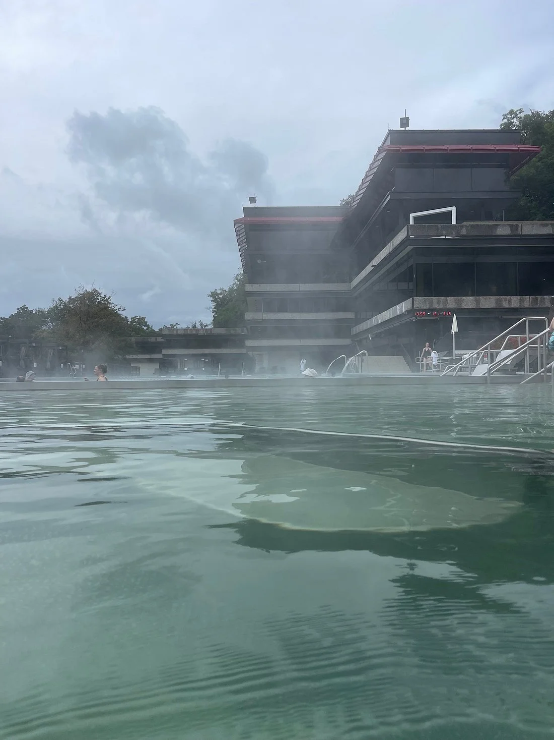 People relaxing in an outdoor swimming pool with a modern building in the background, cloudy sky overhead.