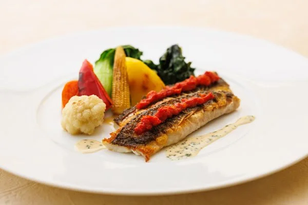 A plated dish with a piece of fish topped with tomato sauce, served with mixed vegetables and a light sauce.