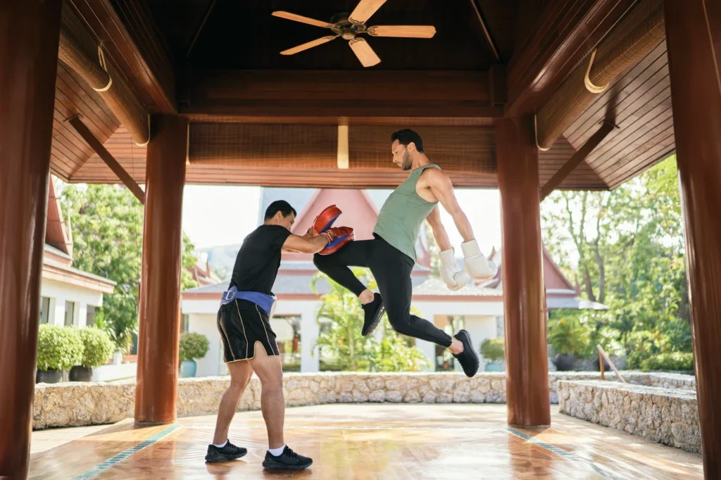 Two men practicing boxing in an outdoor pavilion, one delivering a high kick to the other who is blocking with red focus mitts, with traditional Thai-style roofs and greenery in the background.