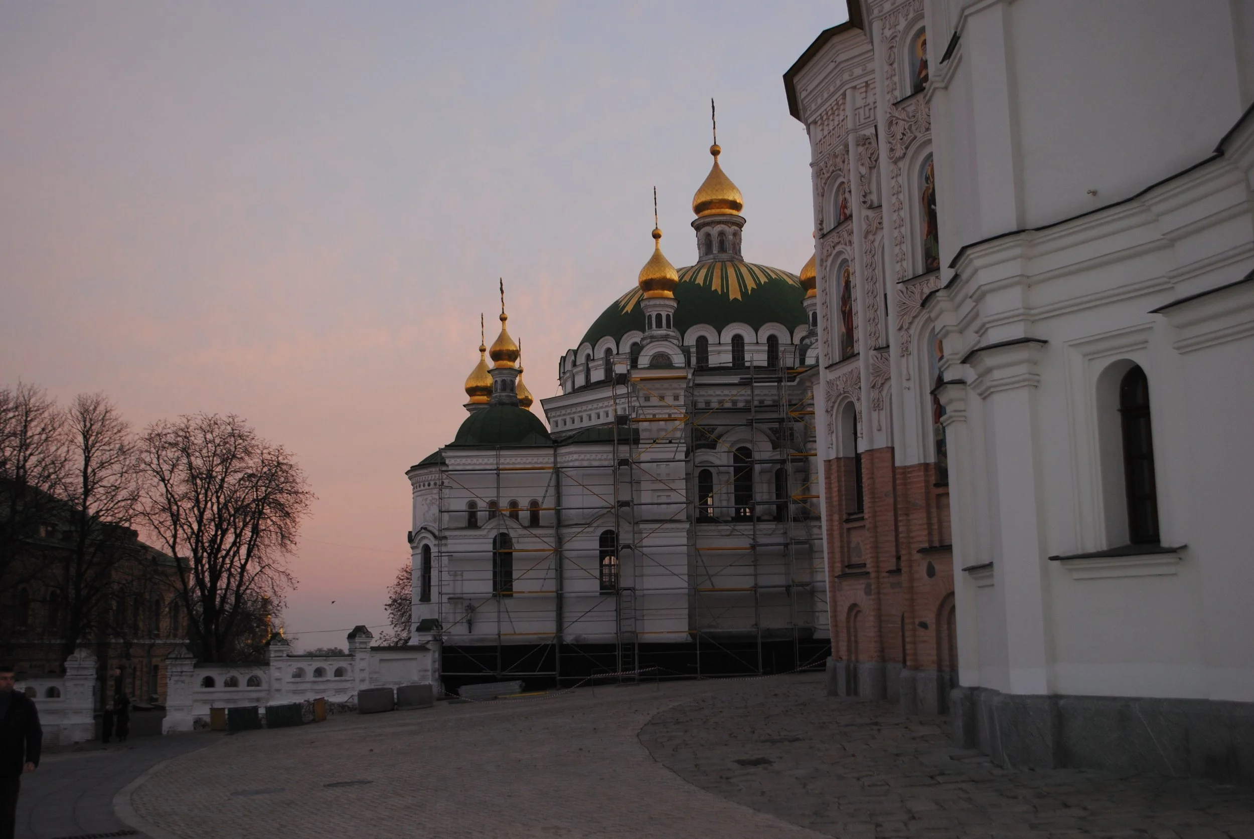 A church with golden domes and onion-shaped spires, surrounded by leafless trees at dusk or early evening.