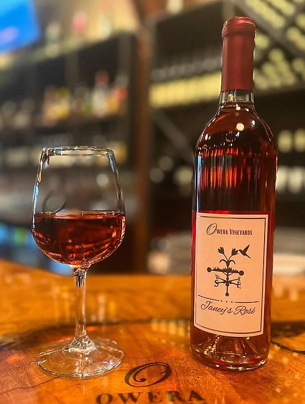 A bottle of Owera Vineyards Janet's Rosé wine and a glass filled with pink rosé wine on a wooden table.
