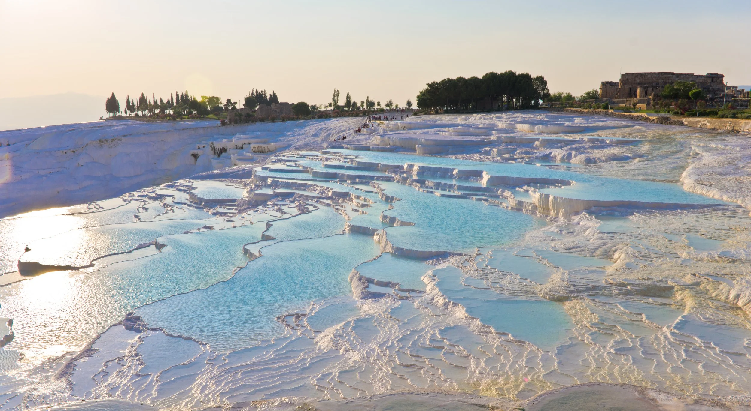Tiered white terraces with blue pools of water, geothermal mineral formations, and a distant tree line at Pamukkale in Turkey during sunset.