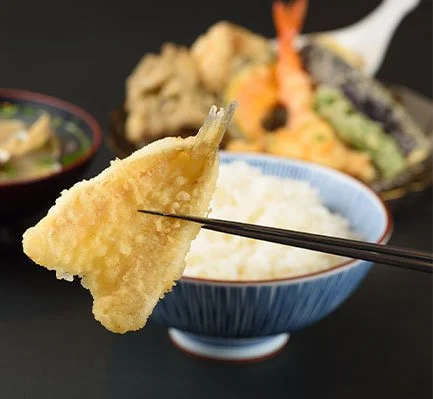 Close-up of a piece of tempura shrimp held with chopsticks over a bowl of white rice, with assorted tempura vegetables in the background.