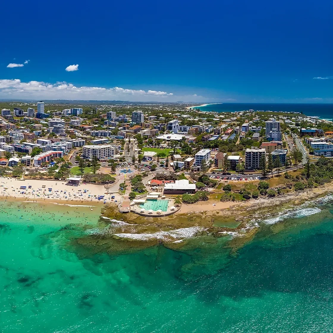 Aerial view of a coastal city with high-rise buildings, a beach with umbrellas, a swimming pool, and the ocean under a clear blue sky.