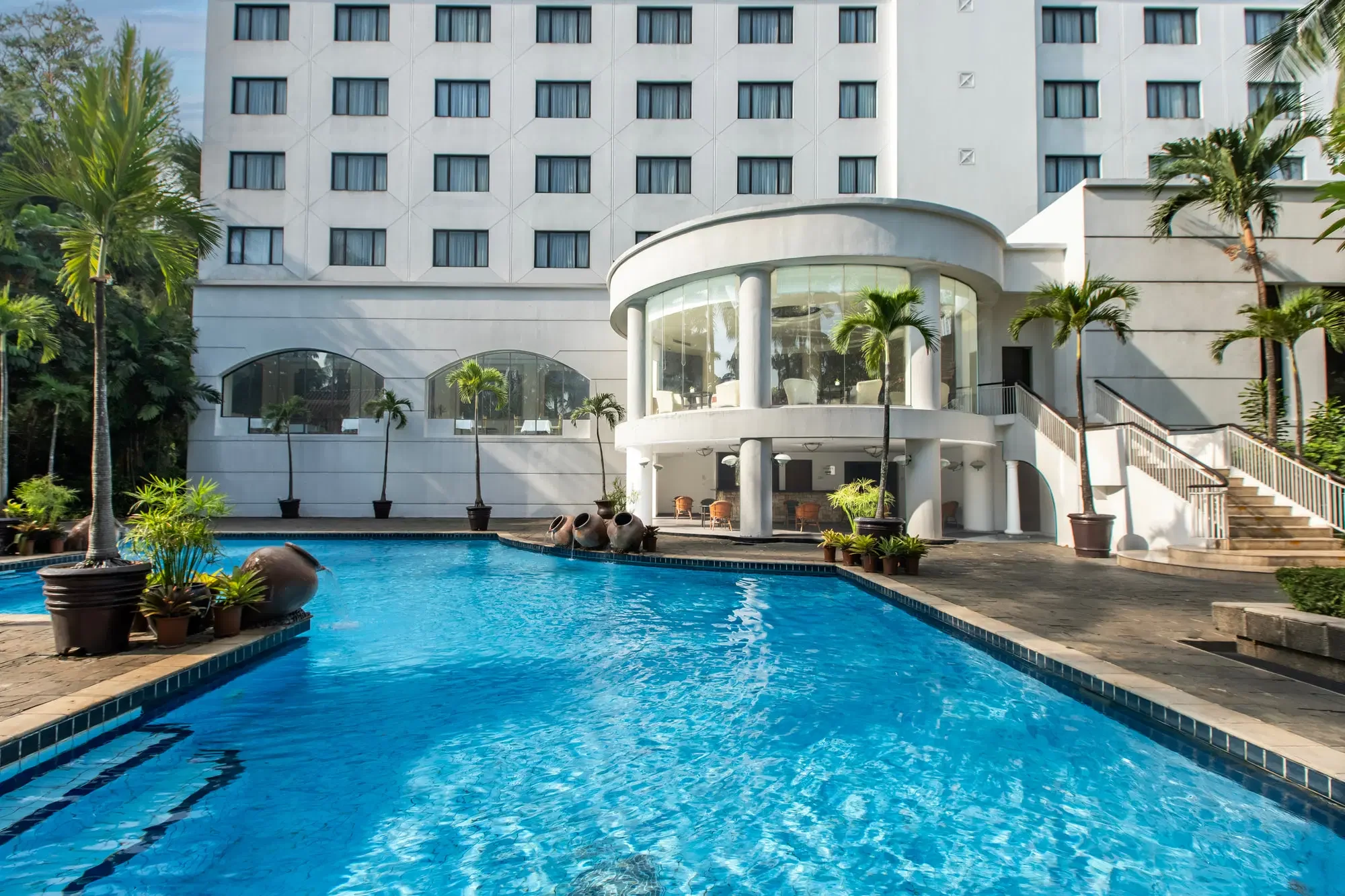 A swimming pool in front of a white multi-story hotel building with multiple windows, tropical plants, and clay pots.