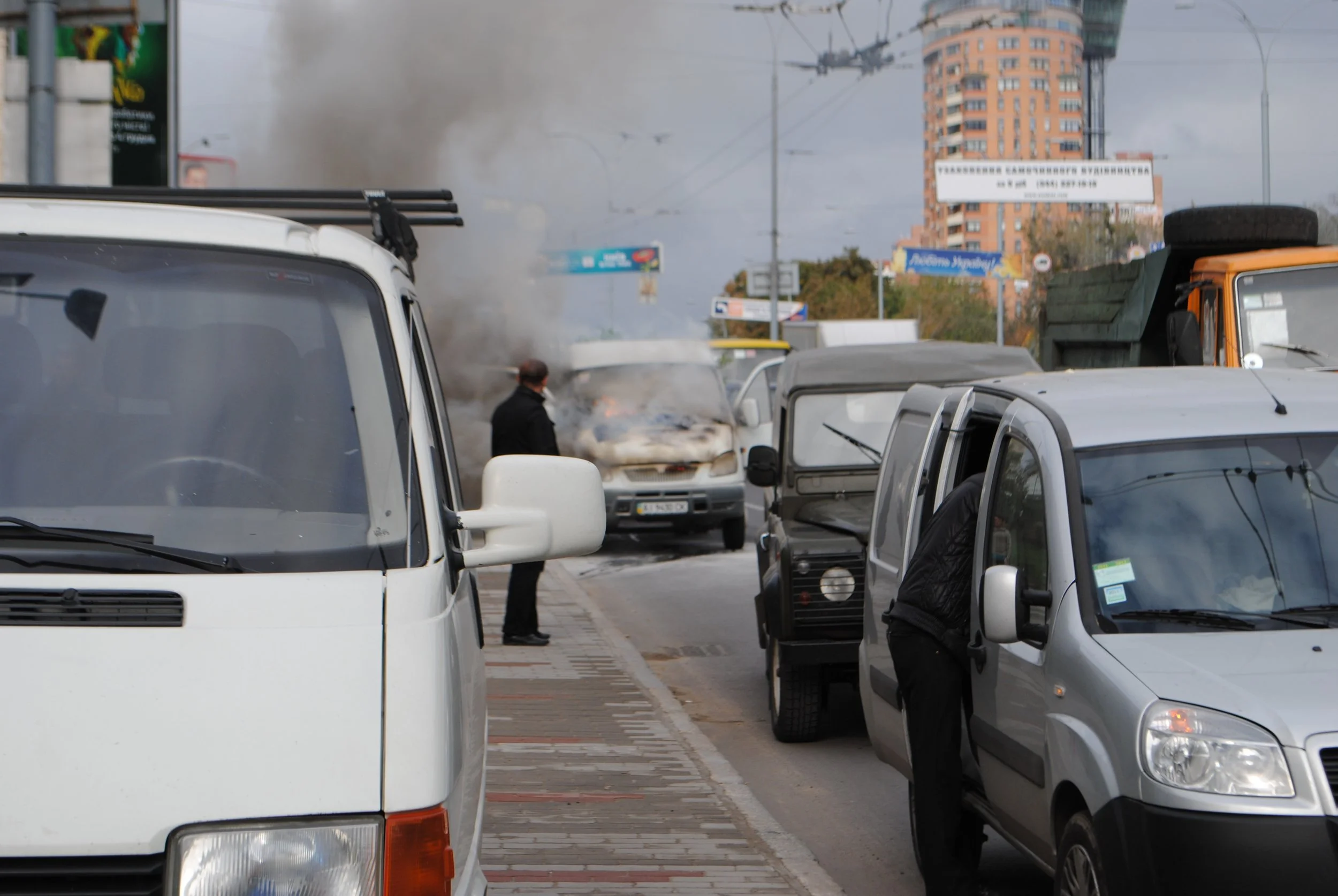 A city street scene showing multiple parked cars, with smoke rising in the background and a person walking among vehicles. Kiev November 2011, Marina Parrish ©