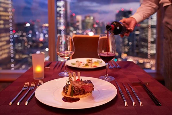 A romantic dinner setup with a plate of steak, two glasses of wine, a candle, and a city skyline view at dusk.