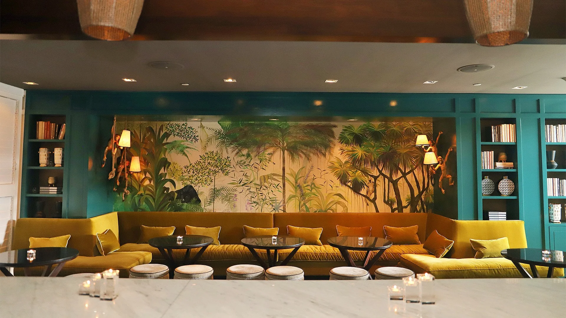 A stylish lounge area with a large green velvet couch decorated with yellow and green pillows, in front of a wall mural of lush green tropical plants and trees, flanked by teal bookshelves filled with books and decorative items, and adorned with hang