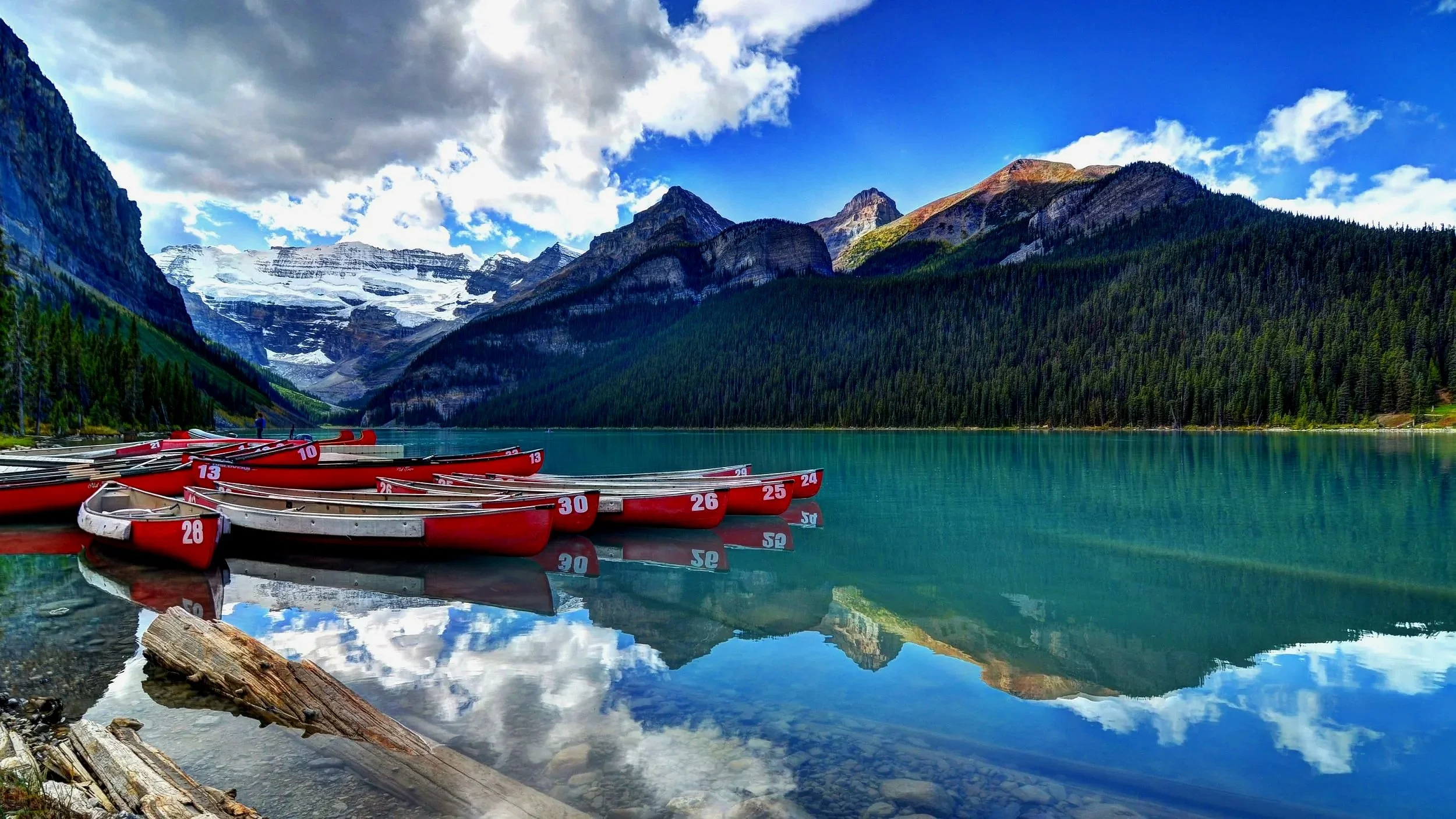 A scenic mountain lake with calm turquoise water, surrounded by lush green pine forests and snow-capped peaks, with a row of red and white canoes docked at the shore.