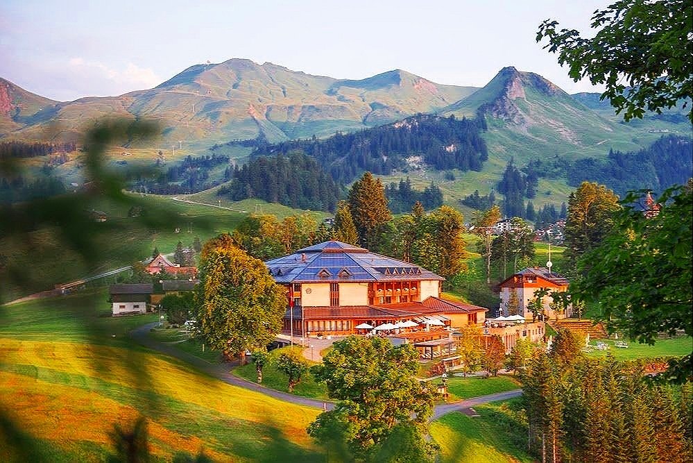 A scenic view of a large building with a blue roof surrounded by trees, rolling green hills, and mountains in the background under a cloudy sky.