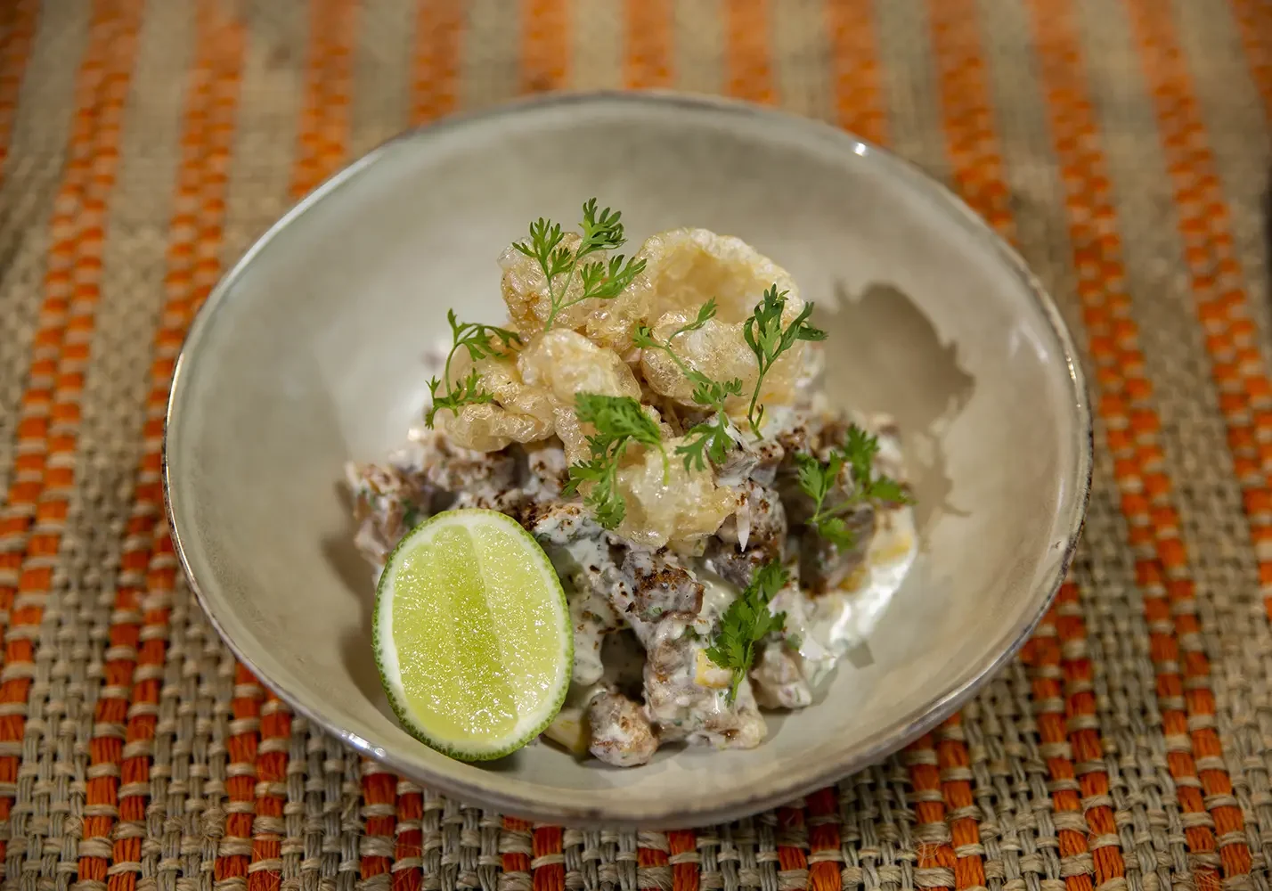 Close-up of a bowl of shrimp cocktail garnished with cilantro, lime wedge, and crispy pork rinds, placed on a woven mat.