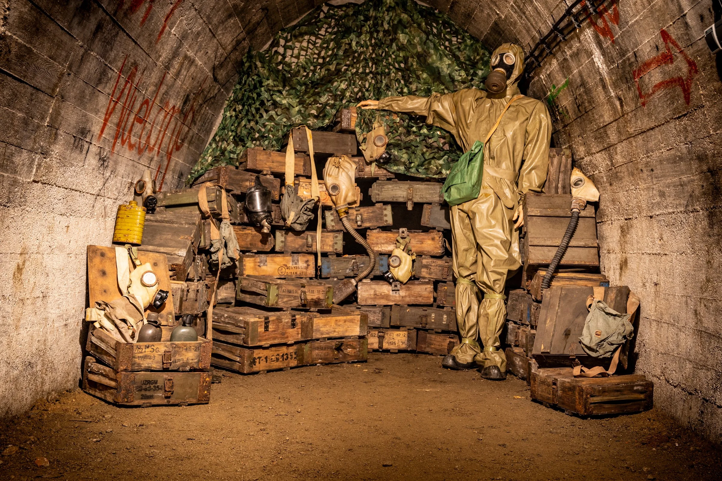 A mannequin wearing a hazmat suit and gas mask standing inside a concrete tunnel. The tunnel is filled with stacked wooden crates, some with gas mask and canister gear hanging on them. Green camouflage netting is draped behind the mannequin, and red 