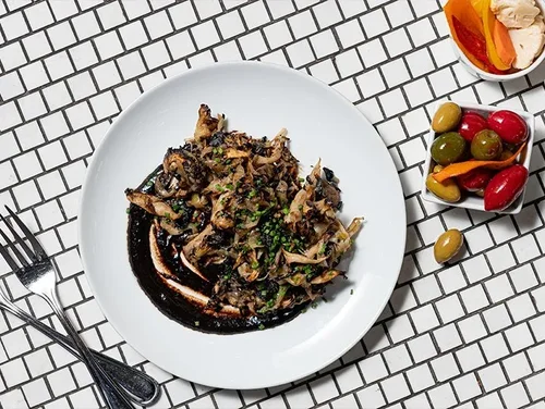 A white plate with shredded meat topped with chopped green herbs, served over a dark sauce on a white tiled table. There are pickles and a small bowl of mixed vegetables nearby, with a fork and knife on the left.