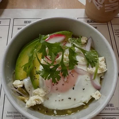 A bowl with a fried egg, avocado slices, radish slices, crumbled cheese, cherry tomatoes, and fresh cilantro on top, with a cup of coffee in the background.