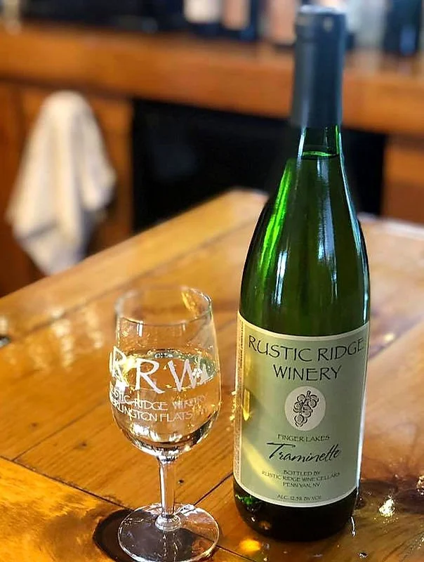 A green wine bottle labeled "Rustic Ridge Winery, Finger Lakes, Traminette" next to a filled wine glass on a wooden table in a cozy setting.