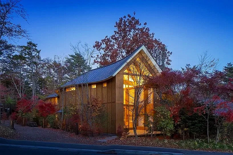 A wooden house with a steeply pitched roof illuminated from inside, surrounded by colorful autumn trees in a wooded area during twilight.