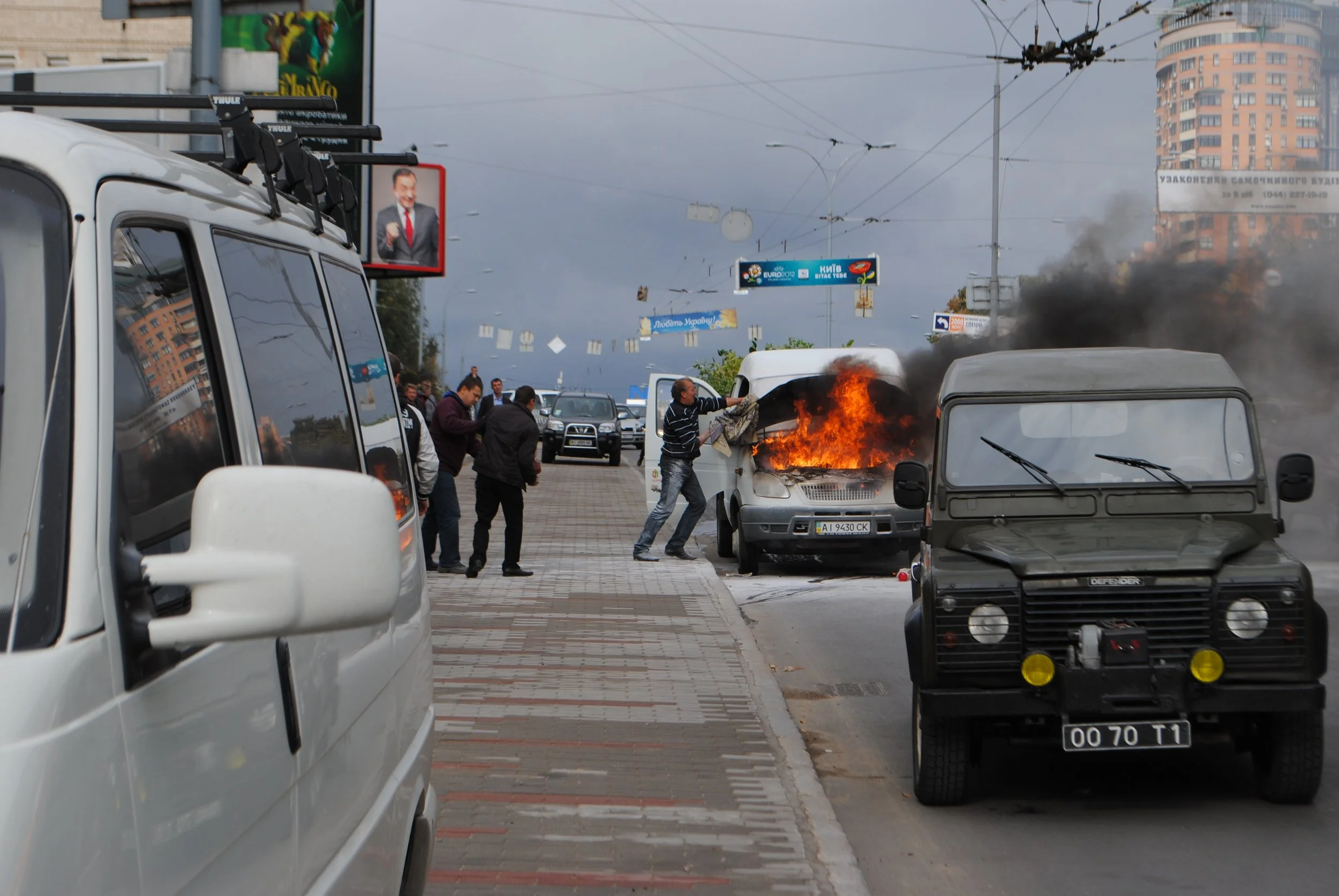 A vehicle fire on a city street with people nearby trying to extinguish the flames, black smoke rising from the car, surrounded by parked and moving vehicles, with buildings and billboards in the background.