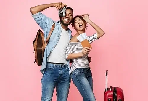 A Cavalier King Charles Spaniel dog laying on a black pet bed with pink piping, on a bed with decorative pillows with colorful patterns, in a room with a dark headboard.