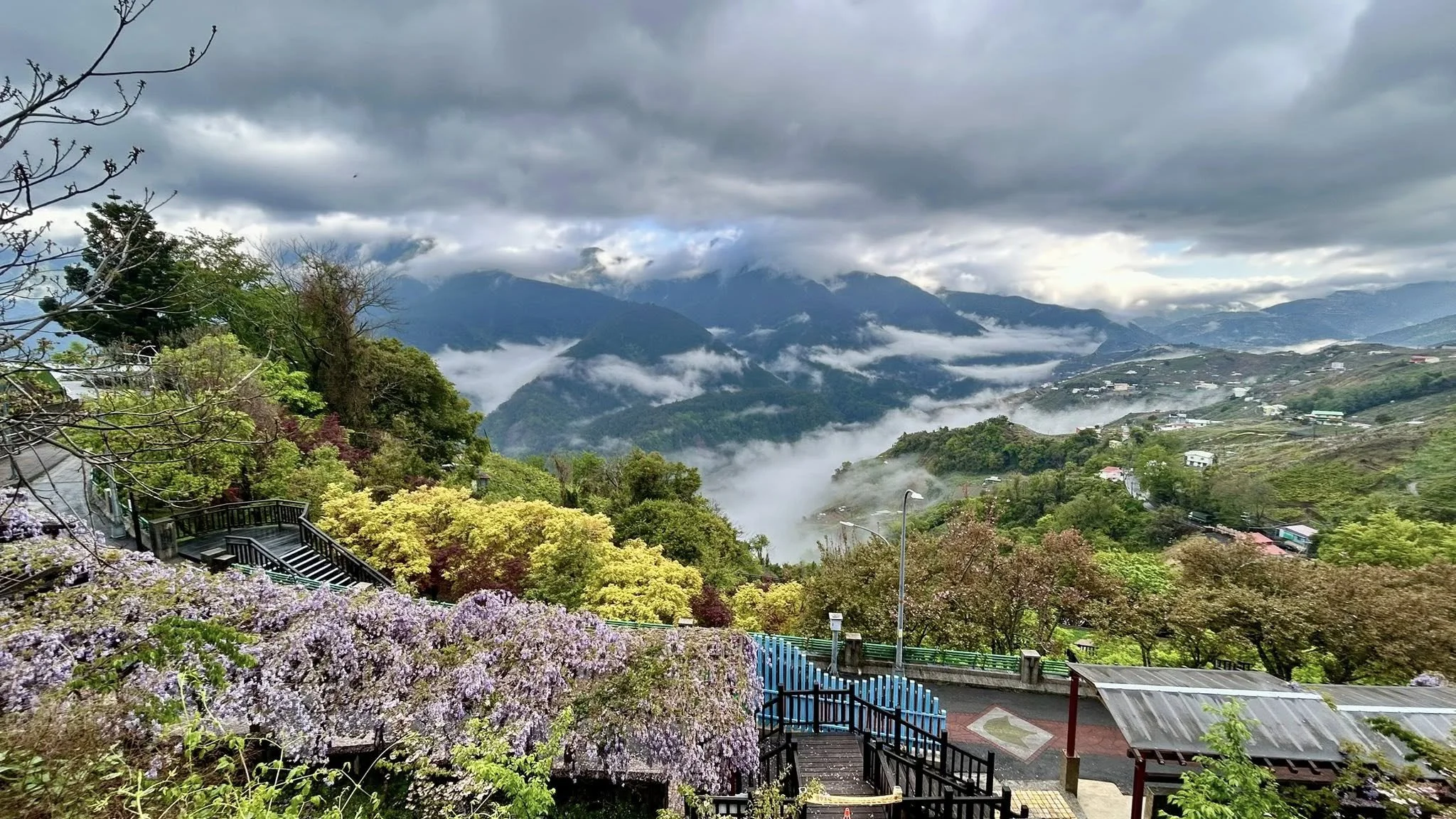 A scenic mountain view with lush green trees and colorful flowers in the foreground. Misty clouds drift through the valleys and partially obscure the mountains in the background, creating a peaceful, misty landscape.