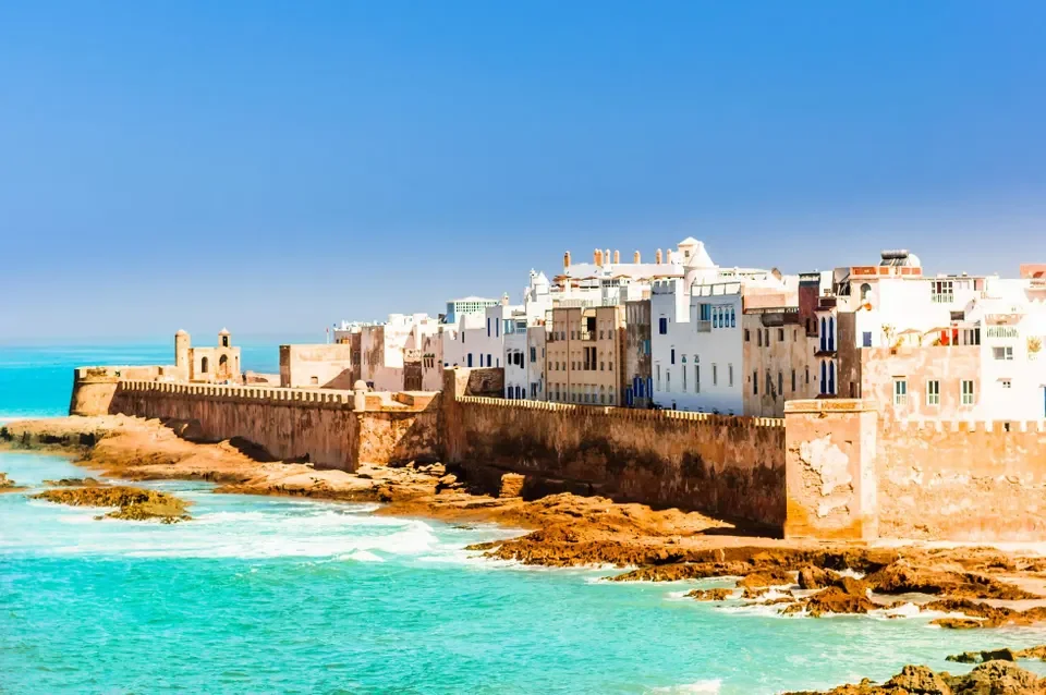 Coastal city with white and beige buildings, a stone wall along the shoreline, turquoise water, and a clear blue sky.