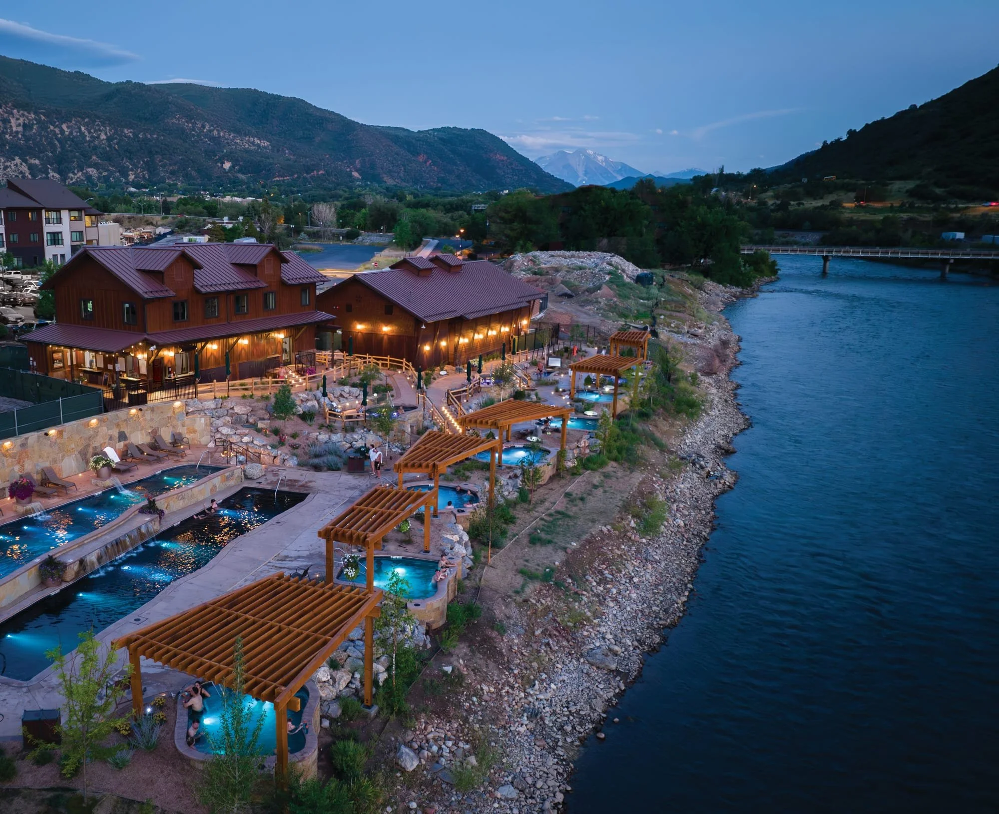 A riverside resort with multiple hot tubs and a wooden building, illuminated by string lights, set in a mountainous landscape during early evening.