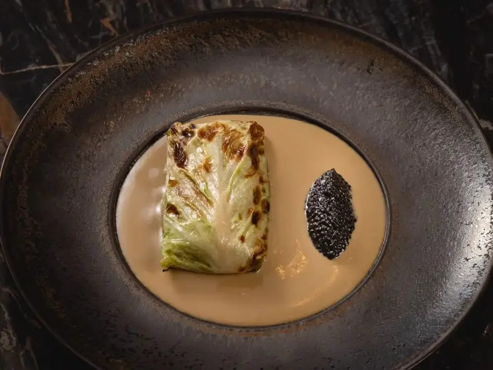 A piece of grilled lettuce in a cream sauce, garnished with a dollop of black caviar, served in a black bowl.