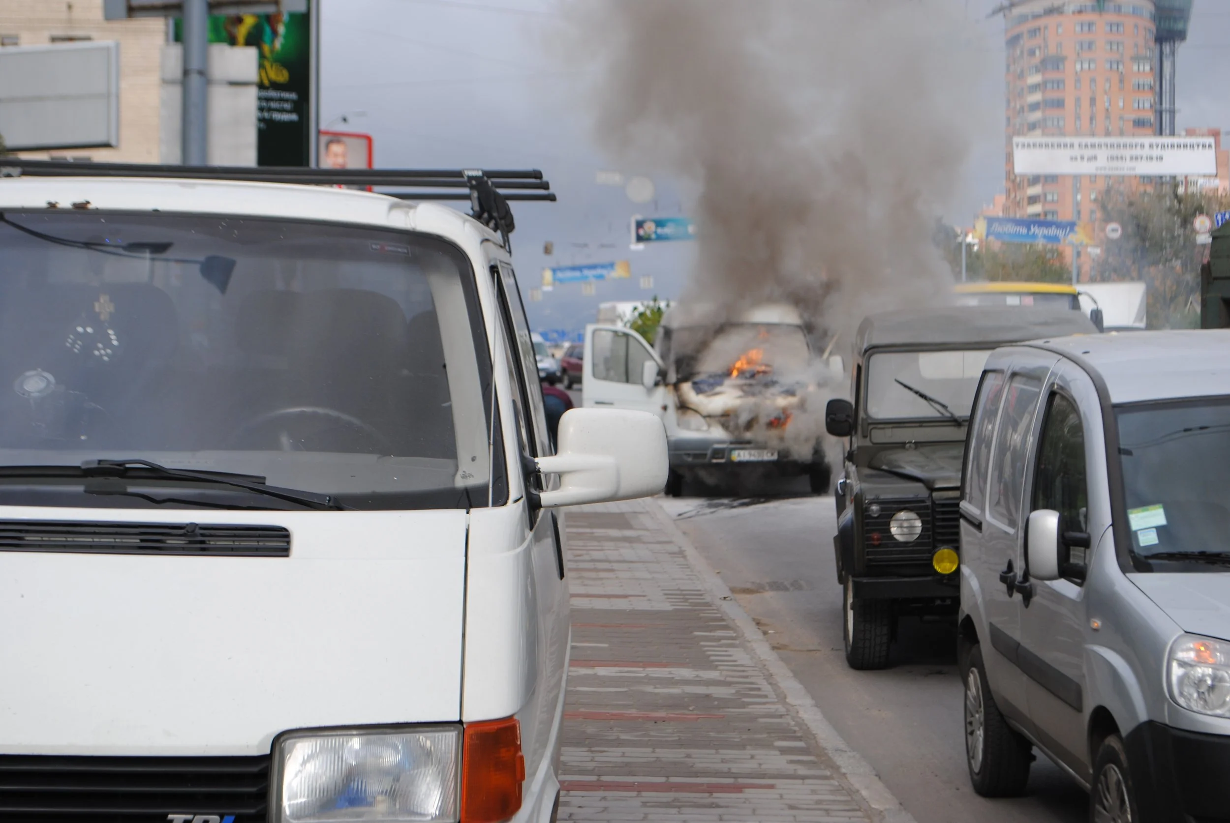 A street scene with a car on fire in the background, thick black smoke rising into the air, multiple parked cars, and buildings with signs in the distance. Kiev November 2011, Marina Parrish ©