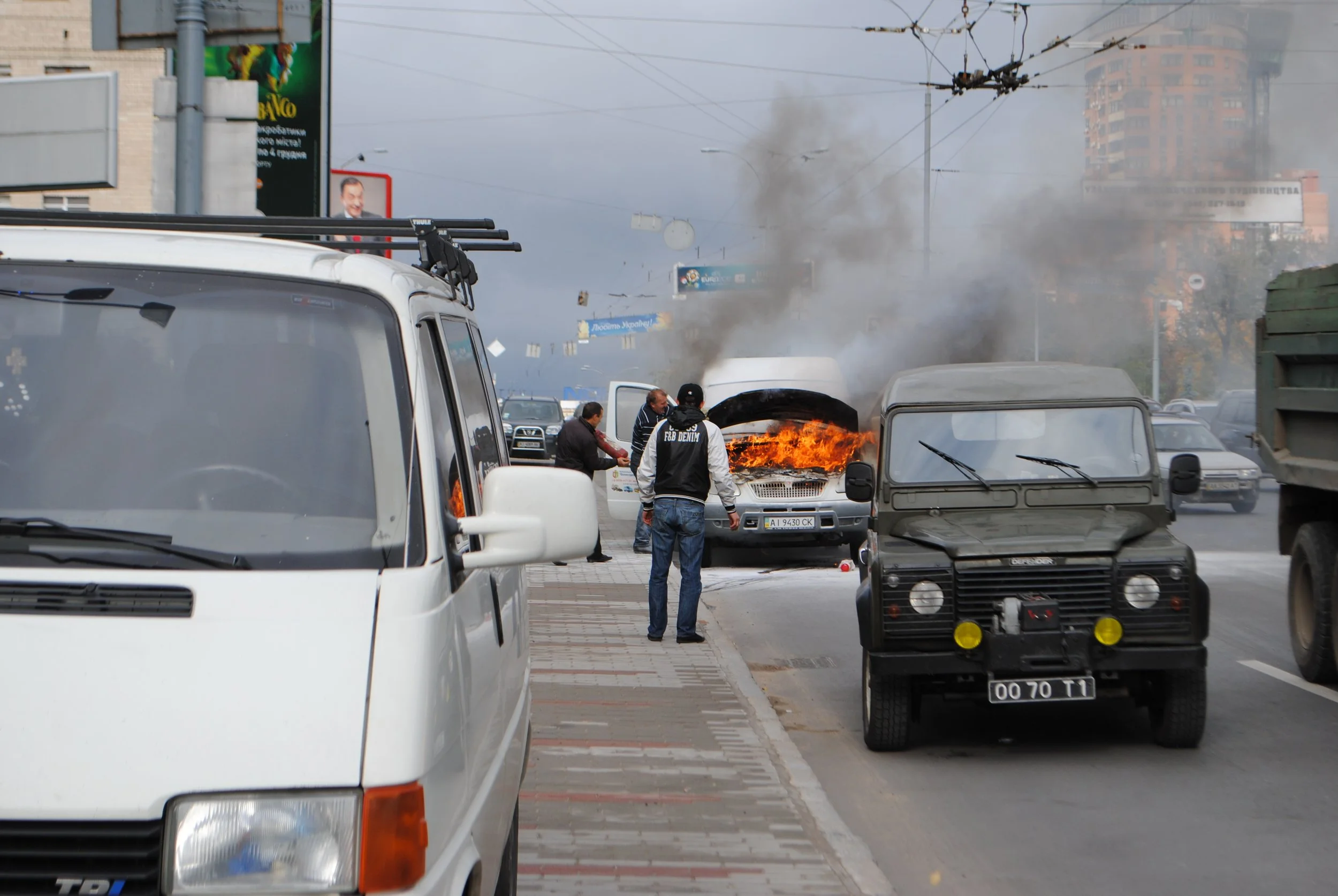 A vehicle on fire with thick black smoke, people standing nearby, and cars parked along the street. Kiev November 2011, Marina Parrish ©