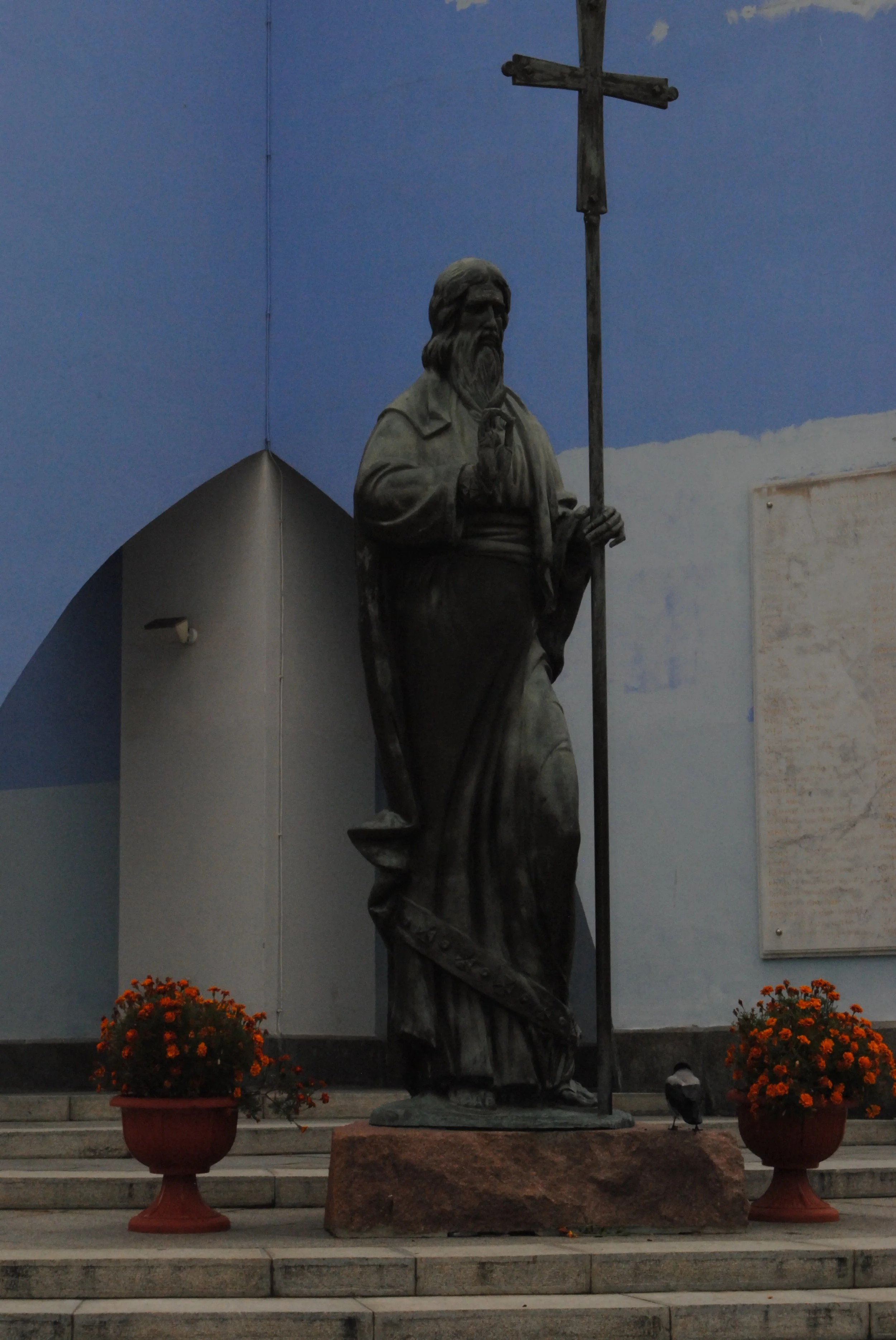 A statue of a religious figure, possibly Jesus or a saint, holding a staff and making a gesture with the right hand,, positioned on a stone pedestal, with two orange flowers in pots on either side, against a blue sky background.
