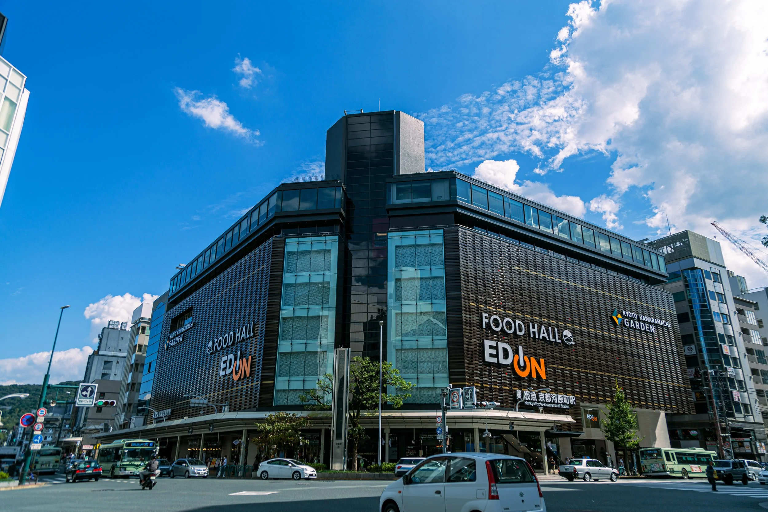 A modern multi-story building with a sign reading 'Food Hall EDUN' and 'Kyoto Kawaramachi Garden' at street level, surrounded by cars and buses under a blue sky with some clouds.